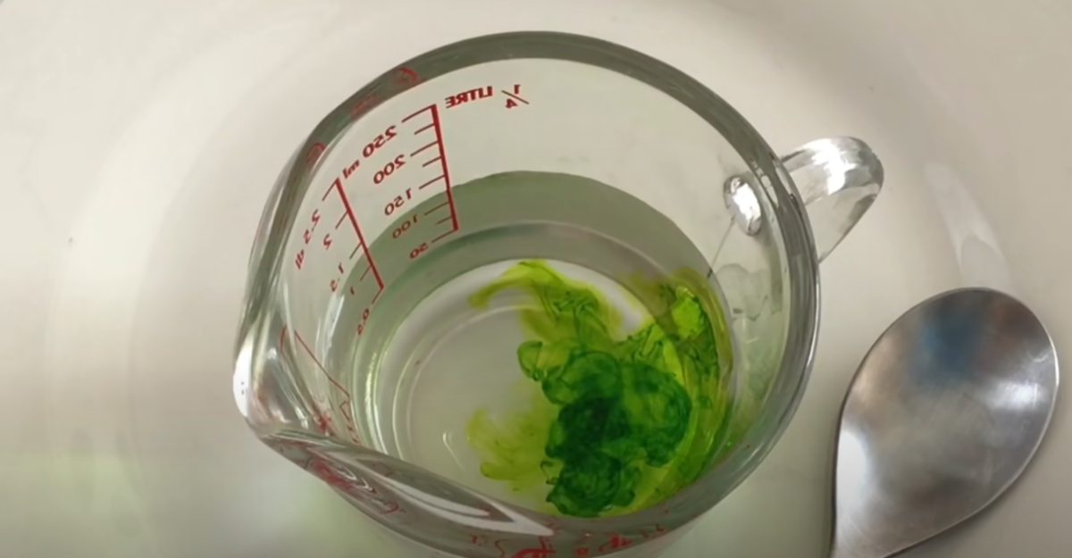 A measuring cup with water in it and a drop of bright green food coloring. 