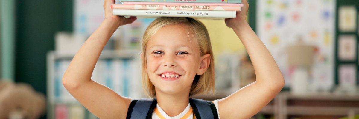 Young girls smiling while holding a stack of books over her head. 