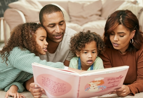 A young family laying on the floor looking at a book together. 