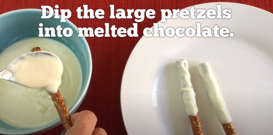 Melted white chocolate and long pretzel rods being dipped into it. 