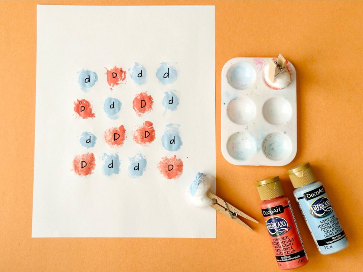 A white paper with lower case letter d's and upper case letter d's that have been painted in blue and orange with a clothespin and cotton ball. 