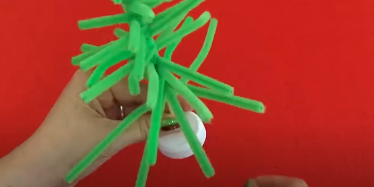 Putting a green pipe cleaner Christmas tree craft into a white plastic cap. 