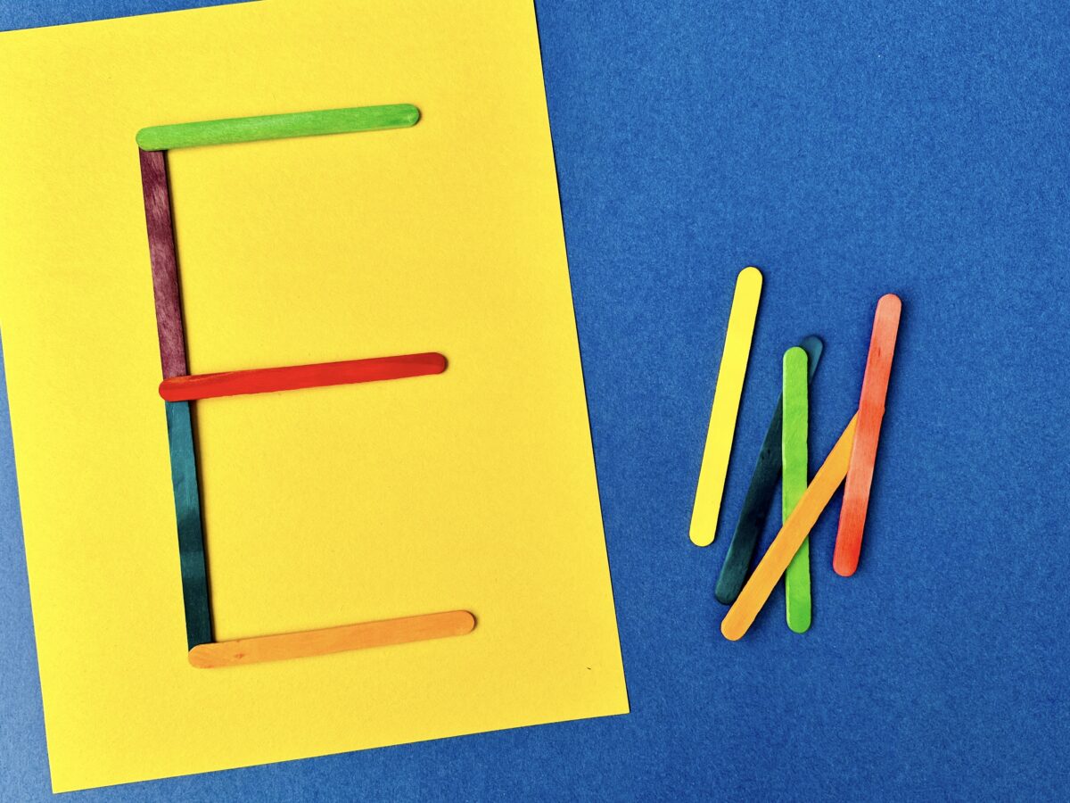 A colorful letter e made from craft sticks on a bright yellow sheet of construction paper. 