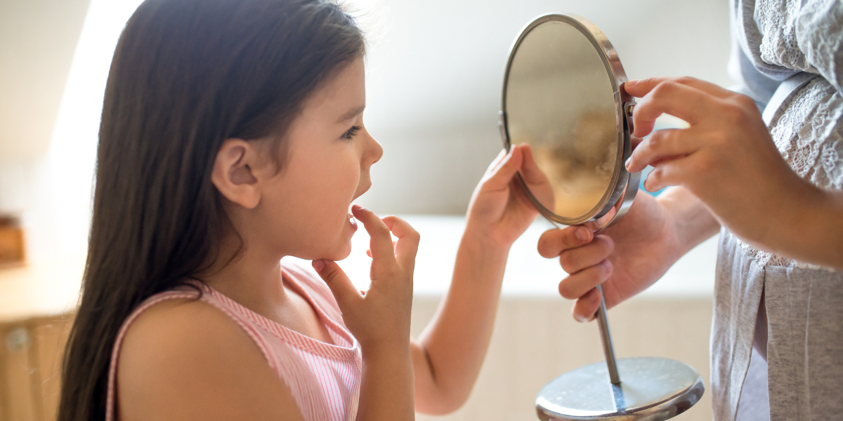 A young girl looks into a hand mirror while pointing at her tongue, observing her mouth or practicing speech.