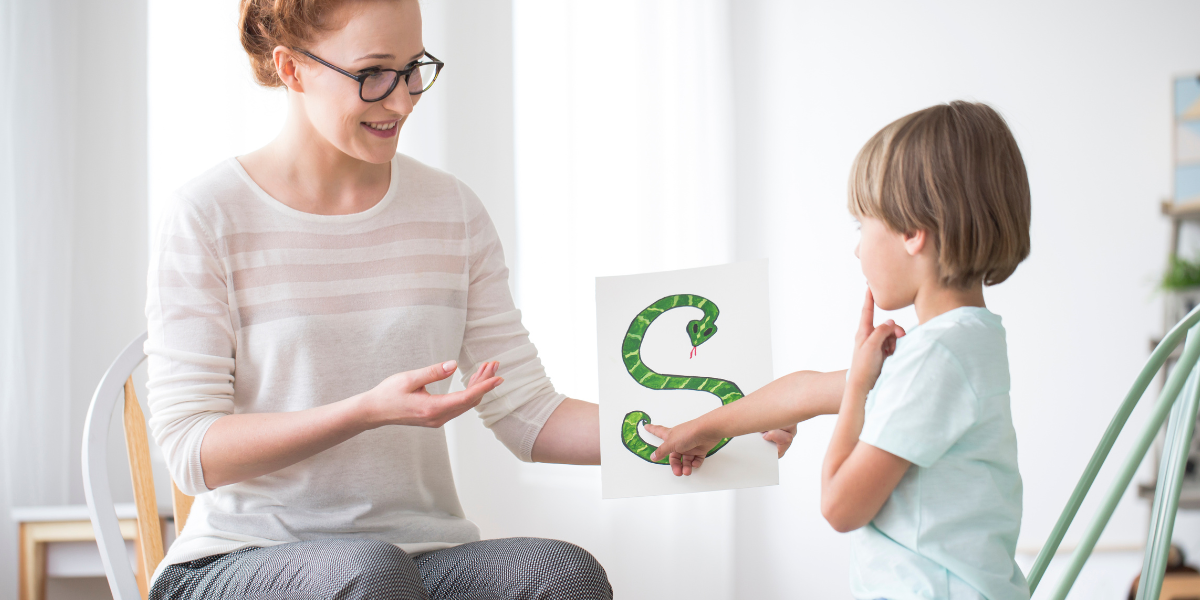 A teacher helps a young boy pronounce the letter 'S' while they both look at a poster of a snake.