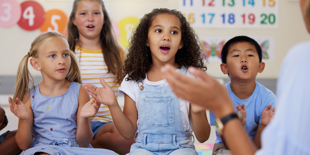 A group of children sitting on the classroom floor, singing and clapping along to a song.