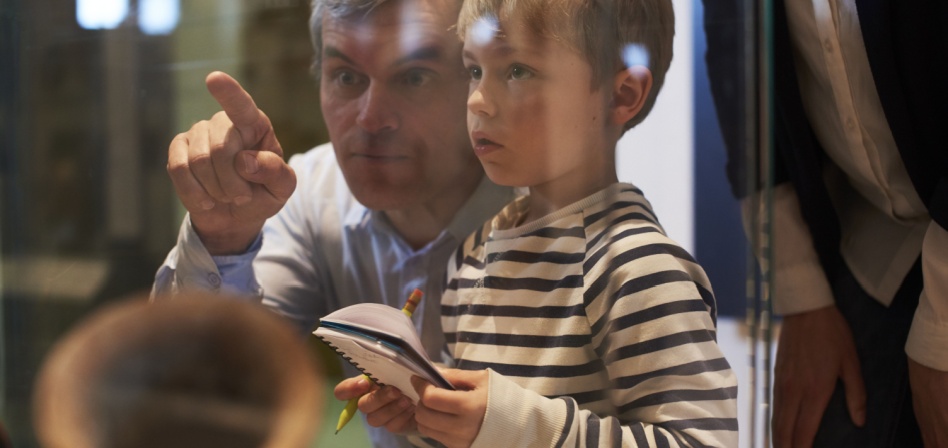 A father pointing out something to his young son who is holding a notebook and a pencil. 