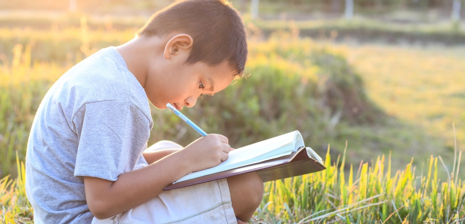 A young boy sitting out side in the setting son writing a description in his notebook. 