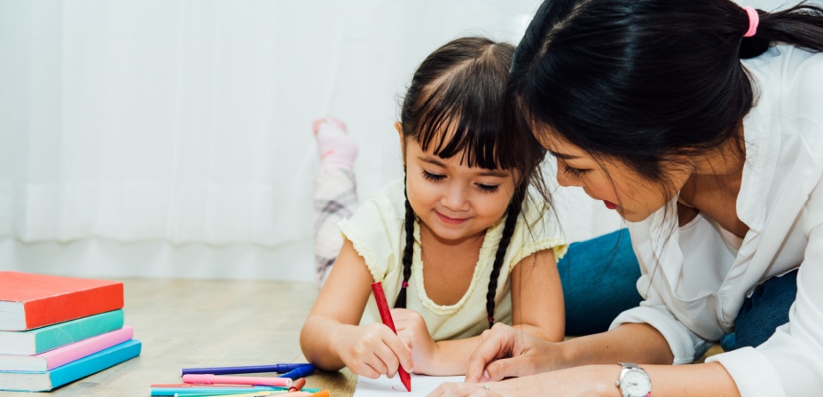 a young girl laying on the floor with her mom drawing in her notebook. 