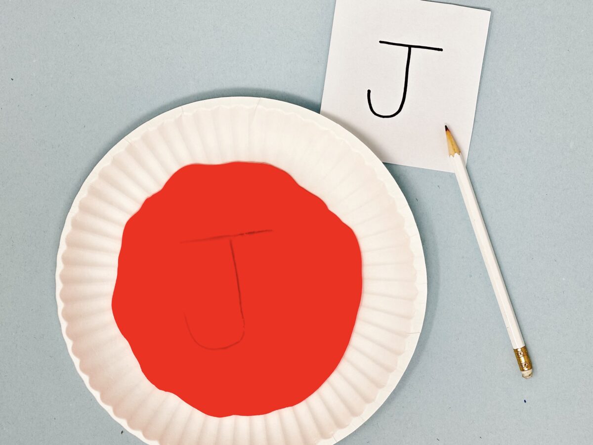 Paper plate filled with red sand featuring the letter J written in the sand. A white piece of paper with a printed letter J is placed beside the plate.