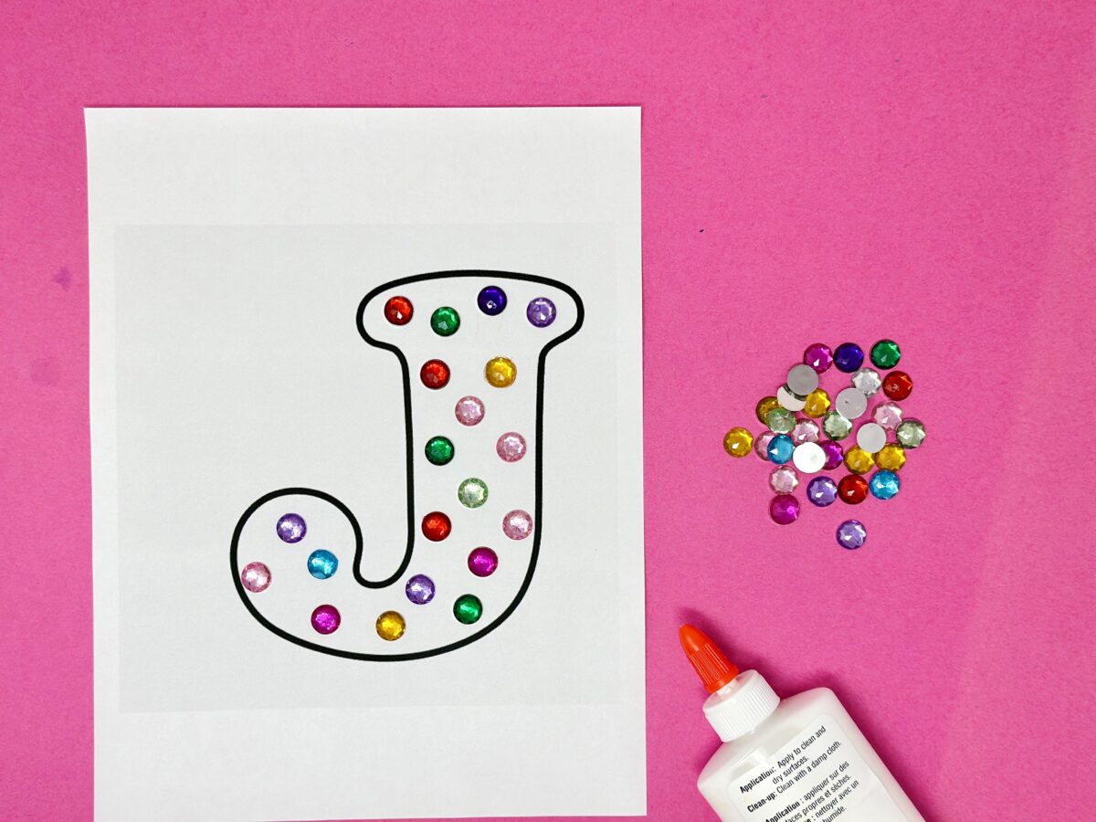 A letter J on white paper covered in colorful jewels. 