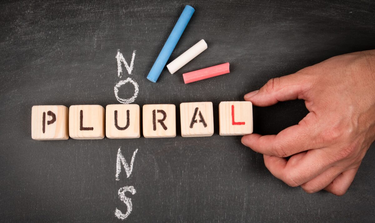 Wood blocks that spell out the word plural, and the word nouns written crosswords style behind it on a chalk board. 