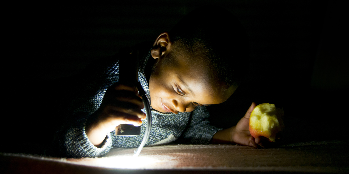 A young child in a dark room with a half eaten apple in one hand, and a flashlight looking down in the other. 