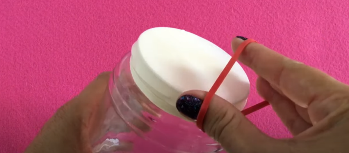 A rubber band being secured over the top of a jar with a white balloon covering it. 