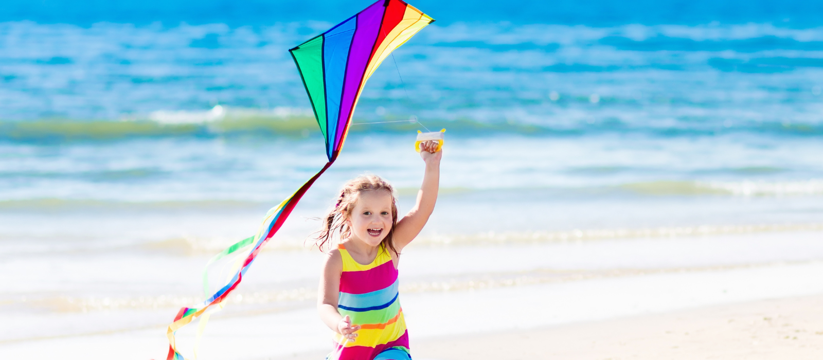 A young girl in a colorful dress playing with a rainbow kite on the beach. 