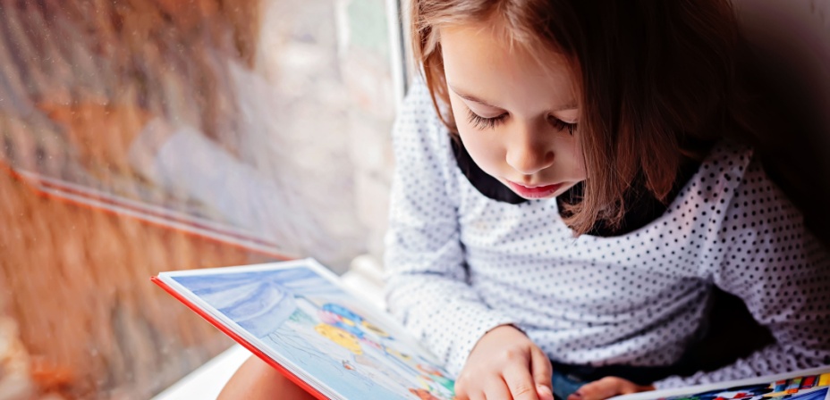 A cute young girl sitting next to a window reading. 