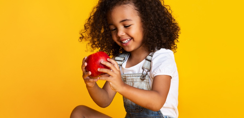 A young girl sitting in front of a yellow backdrop holding a bright red apple and smiling. 