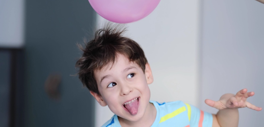 A young boy making a silly face with a pink balloon hovering over his head and making his hair stand on end from the static electricity. 