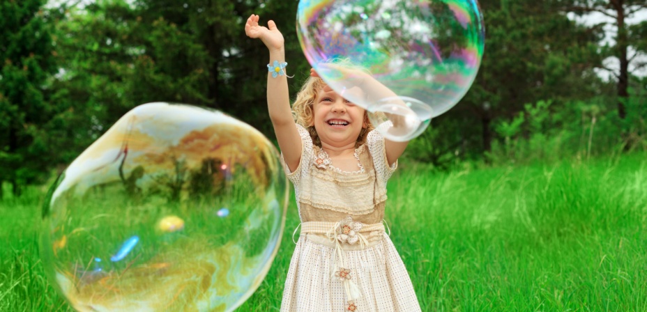 A young girl in a fancy dress outside in a field of long grass watching giant bubbles rise in the air in front of her. 