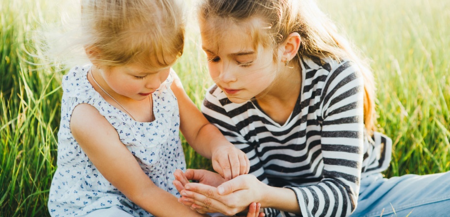 Two young girls sitting in the tall grass. The older girl with hands cusped showing the younger girl an insect she found. 