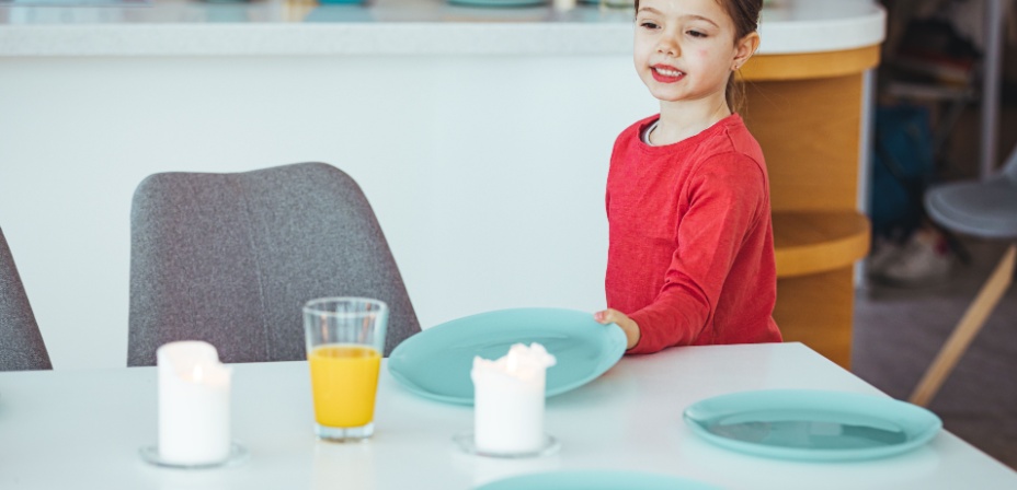 A young girl in a red shirt happily setting the table with plates for dinner. 