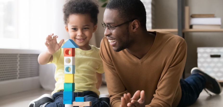 A young father and son laughing and playing together on the floor with wooden blocks. 