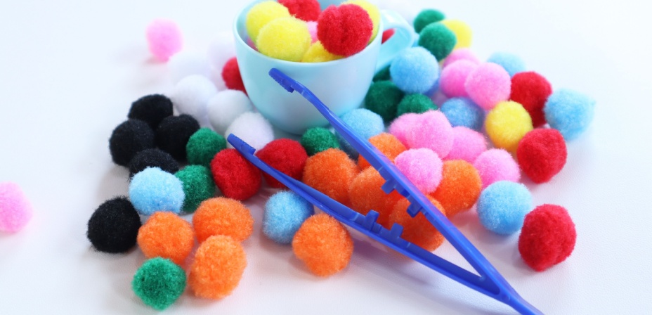 A pile of colorful pompoms and large blue plastic tweezers laying on a white table. 