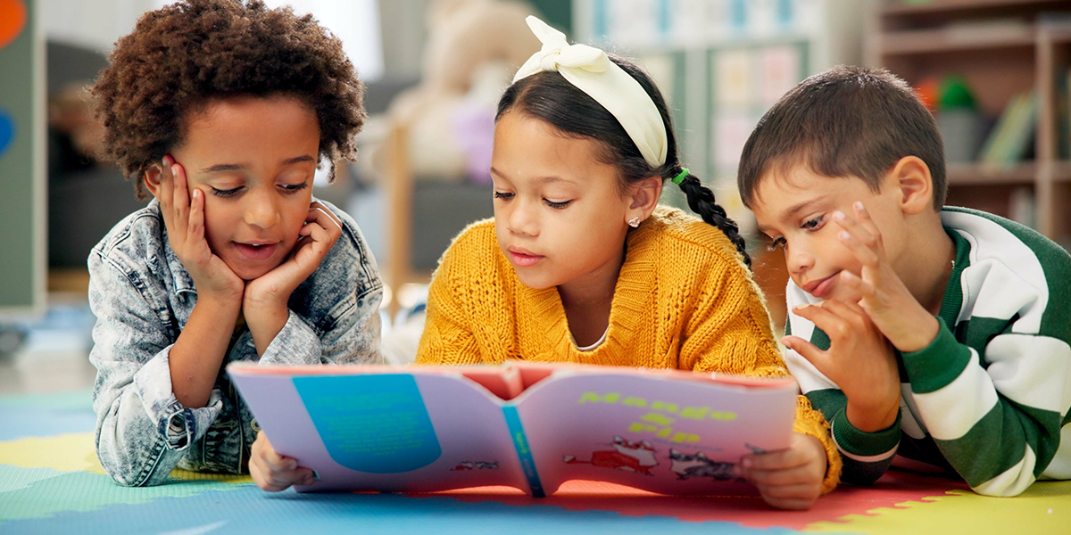 Three children laying on the floor of a library reading a rhyming book. 