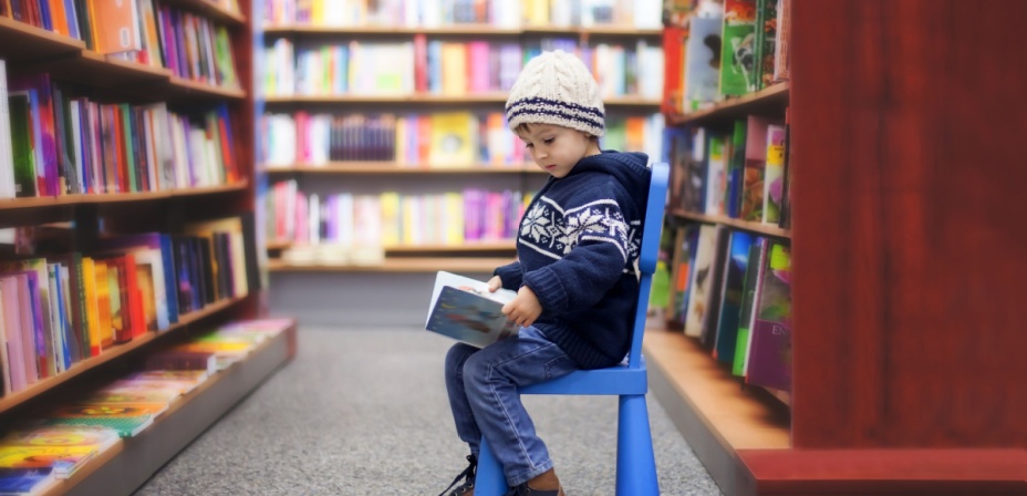 A young boy sitting on a chair in the library reading a book.