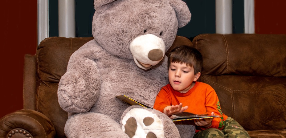 A young boy sitting next to a giant teddy bear reading to it.  