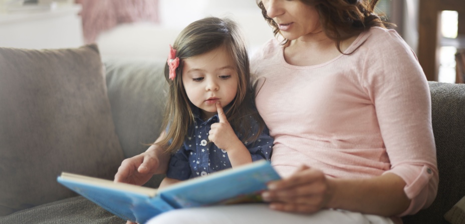 A young mother sitting on the couch with her daughter reading to her. 