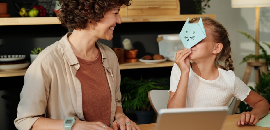 A young girl holding up a folded paper cat face in front of her face while her mom looks at her with a big smile.