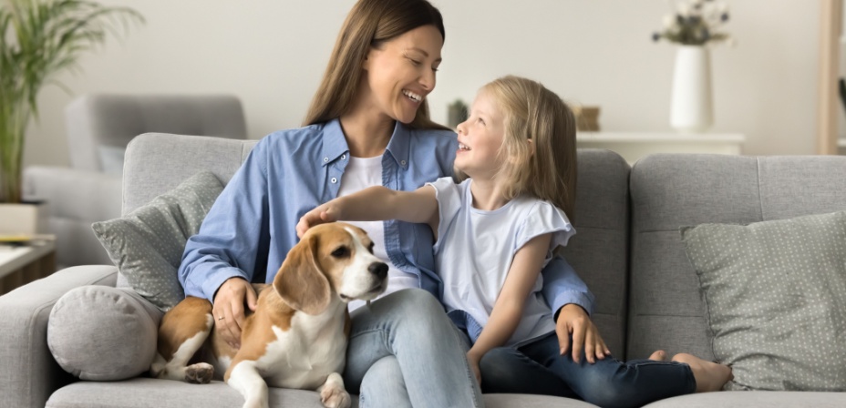 A young mom sitting on the couch with her daughter petting the dog. 