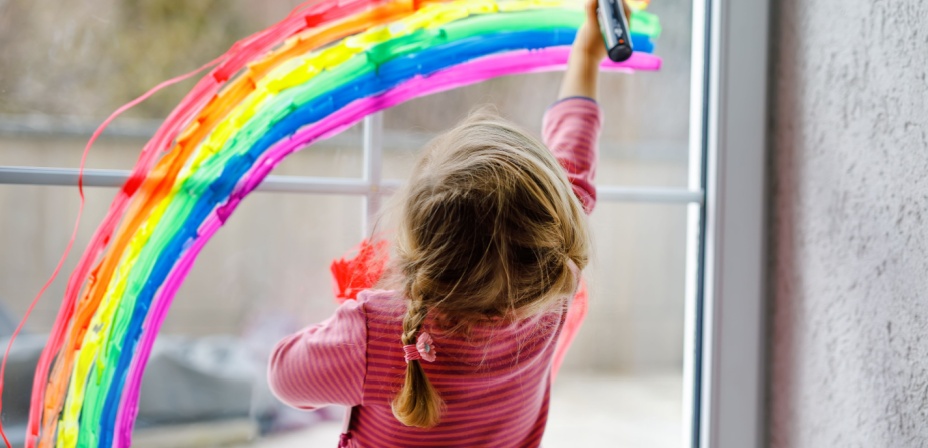 A child drawing a rainbow with window paint on a glass window. 